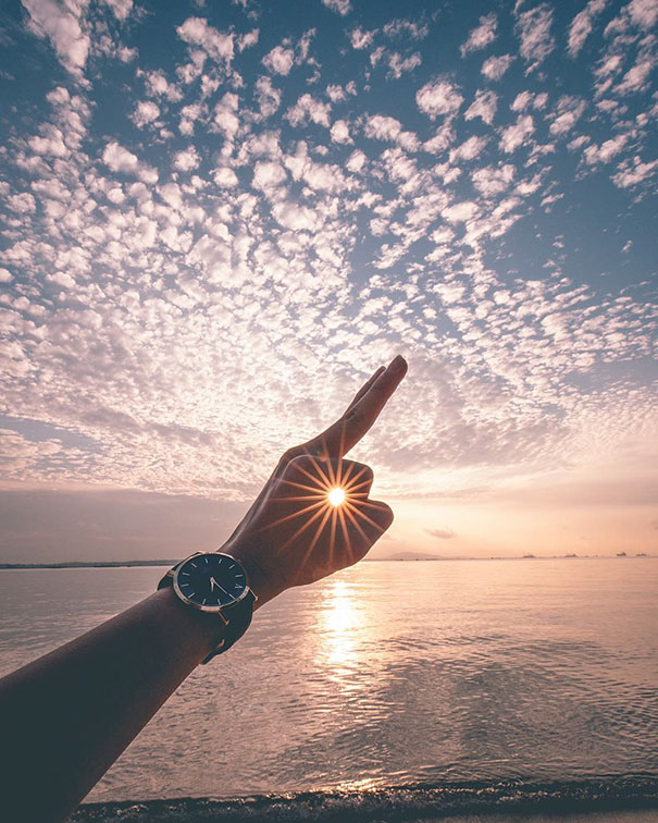 A hand with a watch frames the sun between fingers, creating perfect symmetry over a serene ocean at sunset.