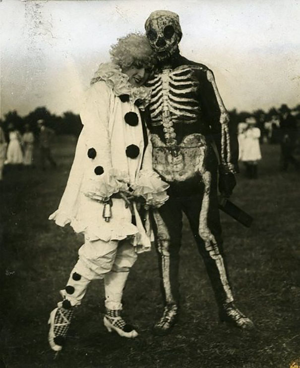 Vintage Halloween costumes featuring a clown and skeleton in a black and white outdoor setting with blurred background figures.