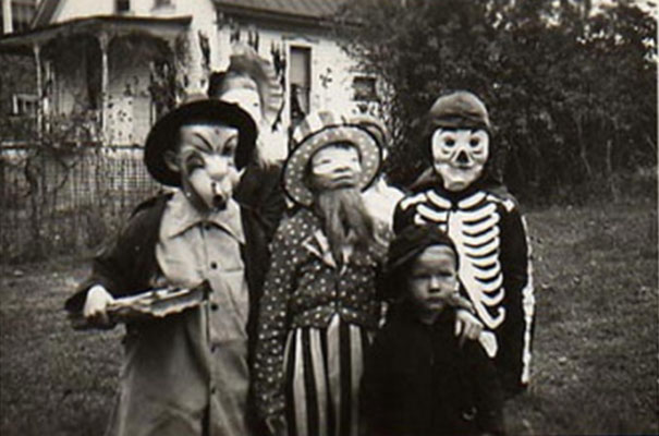 Children in vintage Halloween costumes including a skeleton suit and animal mask posing outdoors in a black and white photo.