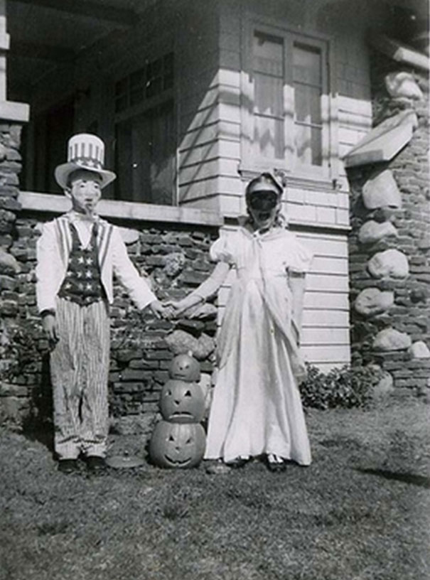 Vintage Halloween costumes featuring children in early 1900s spooky outfits standing by carved pumpkins outside a house.
