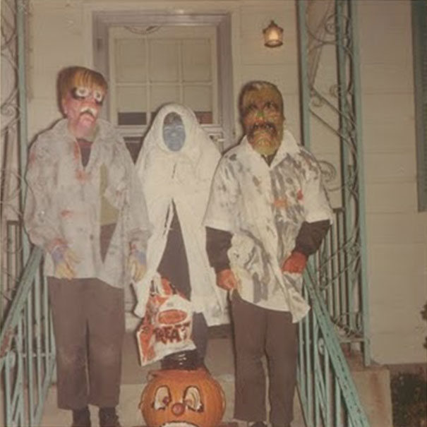 Three people in vintage Halloween costumes with creepy masks standing on a porch holding a treat bag and pumpkin decoration.
