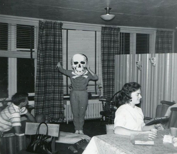 Child wearing a vintage Halloween costume with a large skull mask standing on a chair in a living room.