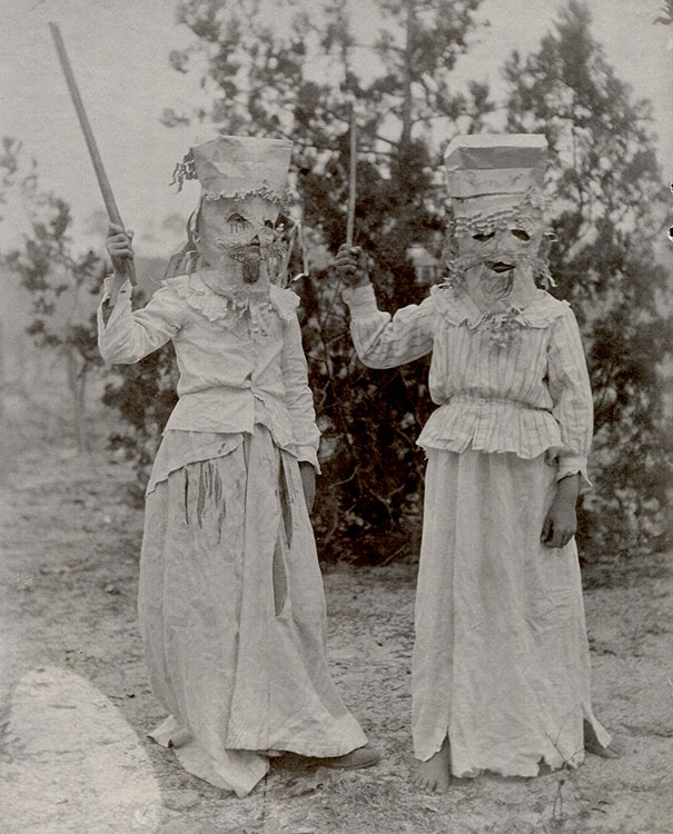 Two children wearing vintage Halloween costumes with paper masks and ragged dresses holding sticks outdoors.