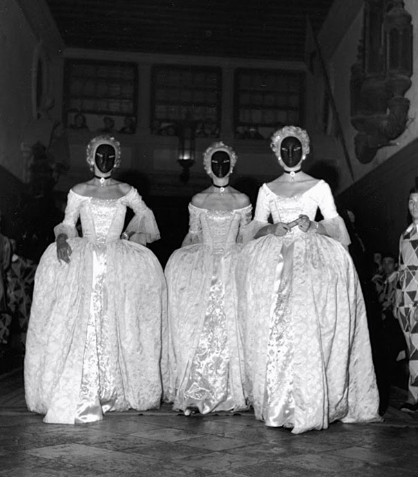 Three people in vintage Halloween costumes wearing elaborate white gowns and black masks in a dimly lit indoor setting.