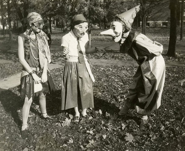 Three children wearing vintage Halloween costumes including masks and a pointed hat, standing outside among fallen leaves.