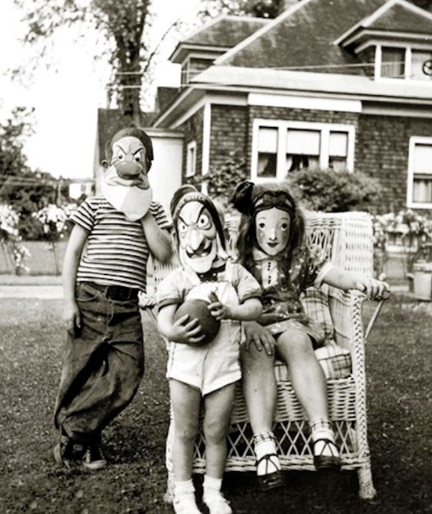 Three children outdoors wearing vintage Halloween costumes and masks, posing in front of a house on a lawn.