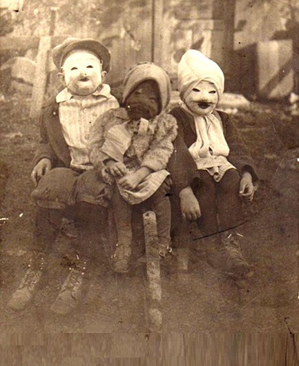 Three children wearing vintage Halloween costumes with eerie masks sitting outdoors in an old sepia photo.