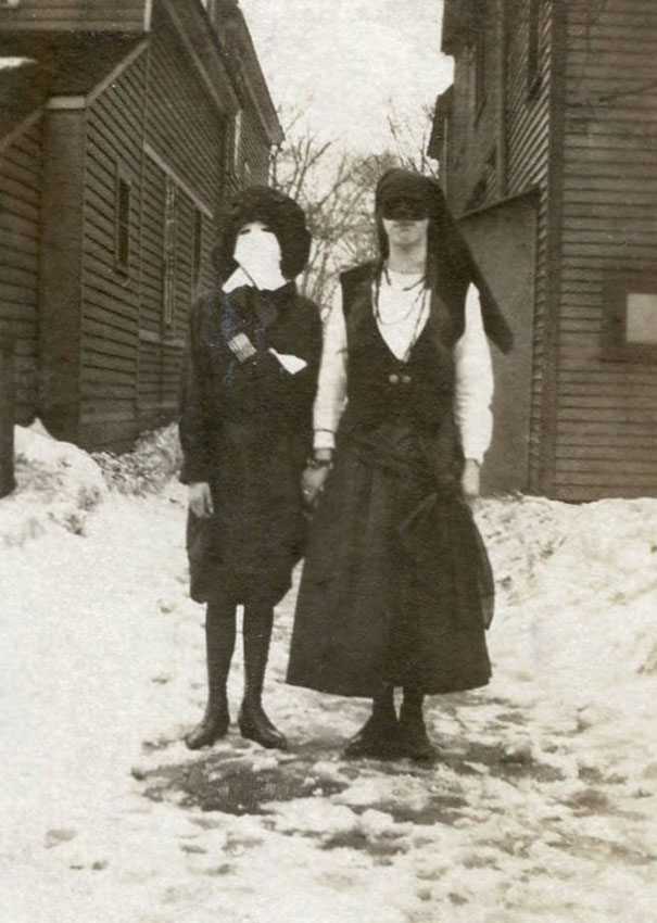 Two people wearing vintage Halloween costumes standing outside between wooden buildings on a snowy ground.