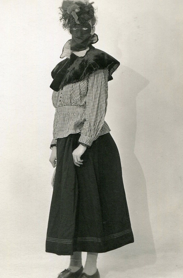 Woman in vintage Halloween costume wearing a mask, checkered blouse, and long dark skirt posing against plain background