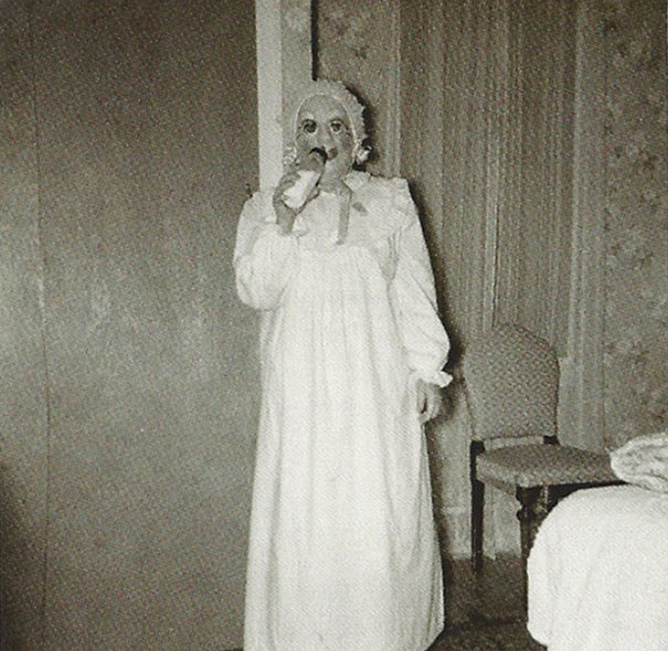 Person wearing a vintage Halloween costume in a long white dress and bonnet with a spooky mask indoors at night.