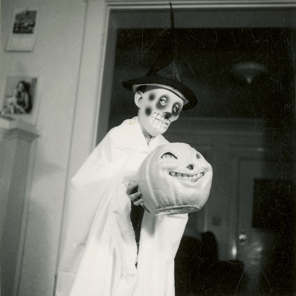 Child in vintage Halloween costume with skeleton face paint and hat holding a grinning pumpkin prop indoors.