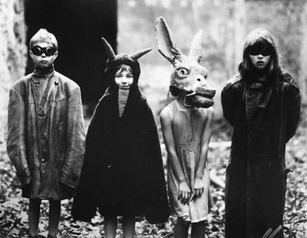 Four children wearing vintage Halloween costumes including a horned cloak, animal mask, and dark glasses in an outdoor setting.
