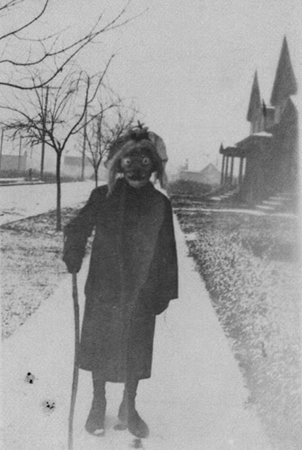 Child in a vintage Halloween costume wearing a mask and coat, holding a stick, standing on a sidewalk beside bare trees.