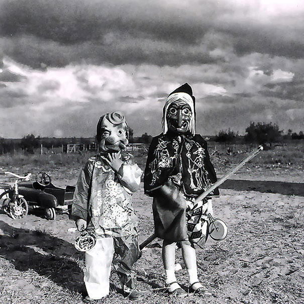 Two children wearing vintage Halloween costumes with creepy masks outdoors on a cloudy day in a rural setting.