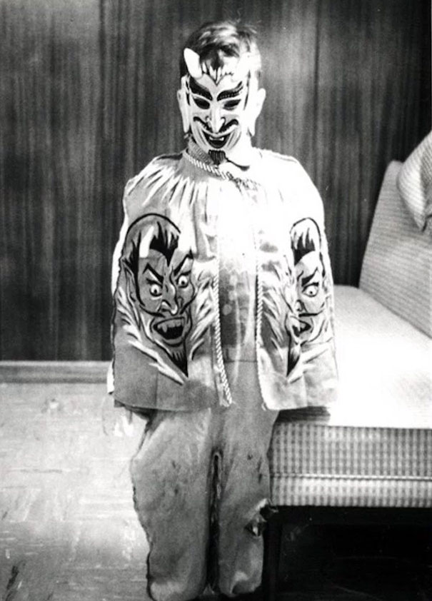 Child in vintage Halloween costume with devil mask and printed cape featuring scary faces indoors, black and white photo