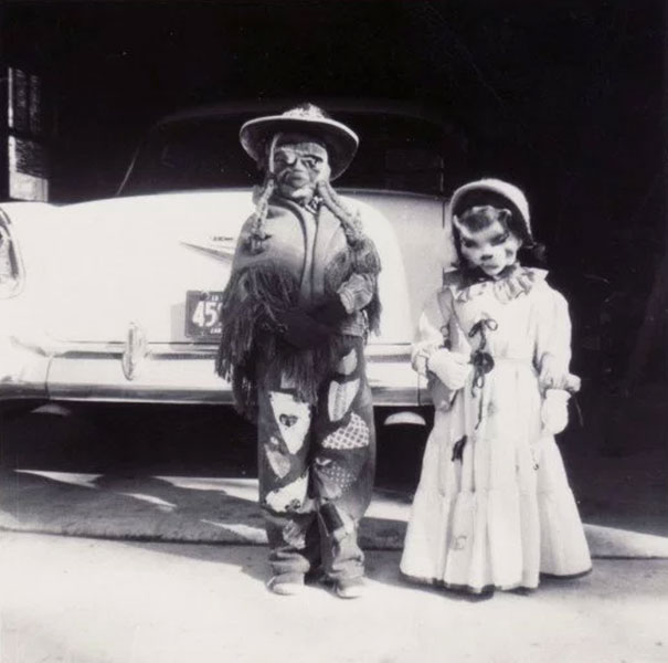 Two children in vintage Halloween costumes standing in front of a classic car, evoking eerie and scary Halloween vibes.