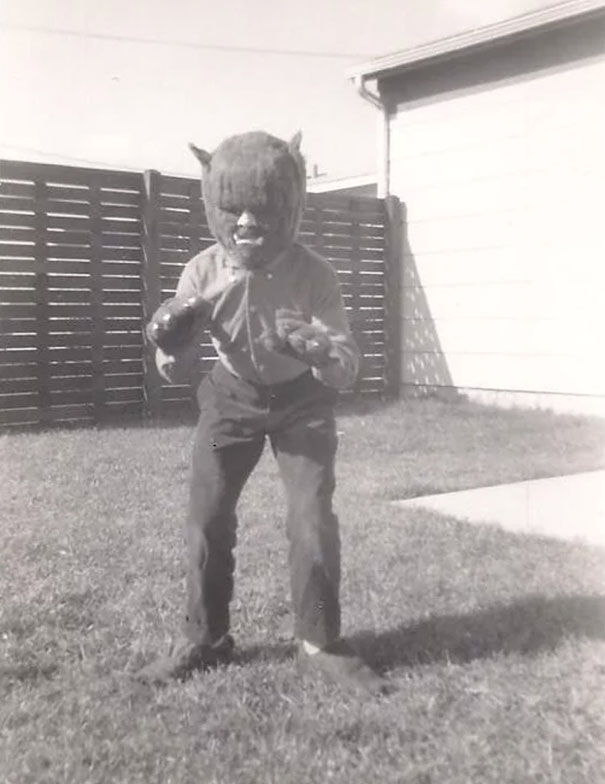 Child in a vintage Halloween costume wearing a werewolf mask and gloves standing in a backyard in black and white.