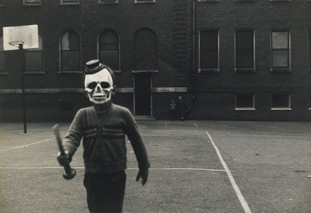 Child wearing a vintage Halloween costume with a skull mask and hat, holding a stick outside a brick building.