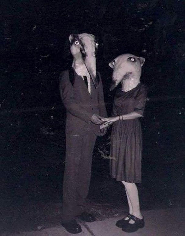 Two people in vintage Halloween costumes wearing large animal-like masks, holding hands in a dark setting.