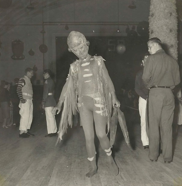 Person in a vintage Halloween costume resembling a skeleton with torn fabric, posing in a dimly lit room with others nearby.