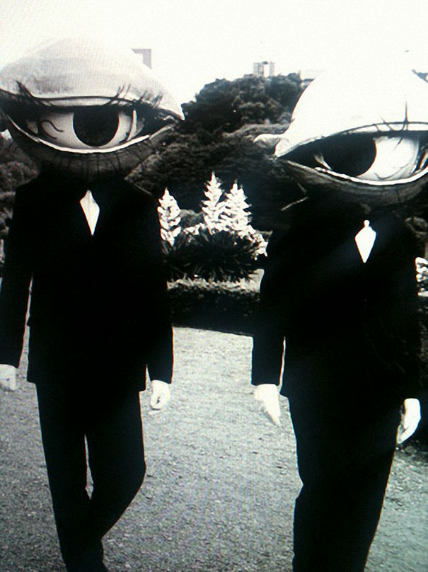 Two people wearing vintage Halloween costumes with large eyeball heads and black suits in an outdoor setting.