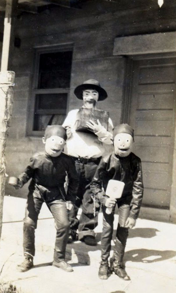 Three people wearing vintage Halloween costumes with homemade masks and simple props standing on a porch in black and white.