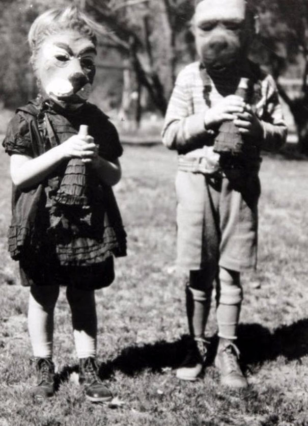 Two children in vintage Halloween costumes wearing animal masks and holding treat bags outdoors on a sunny day.