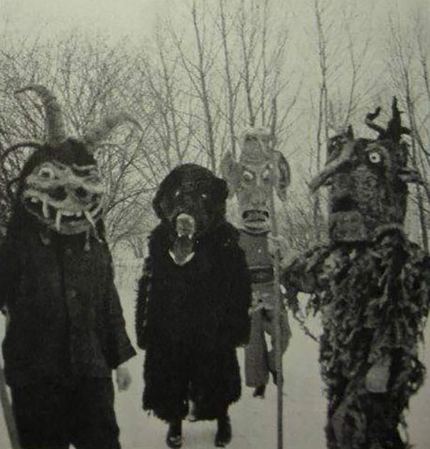 Four people outdoors in vintage Halloween costumes with eerie masks and dark outfits among leafless trees.