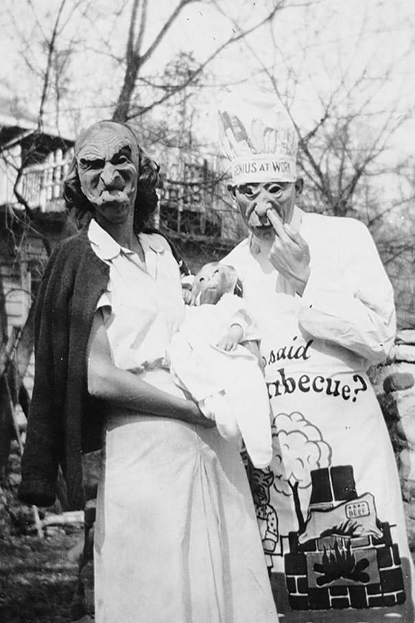 Two people in vintage Halloween costumes with creepy masks holding a baby doll outdoors in a black and white photo.