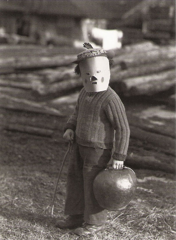 Child in a vintage Halloween costume wearing a homemade mask and holding a pumpkin outdoors.