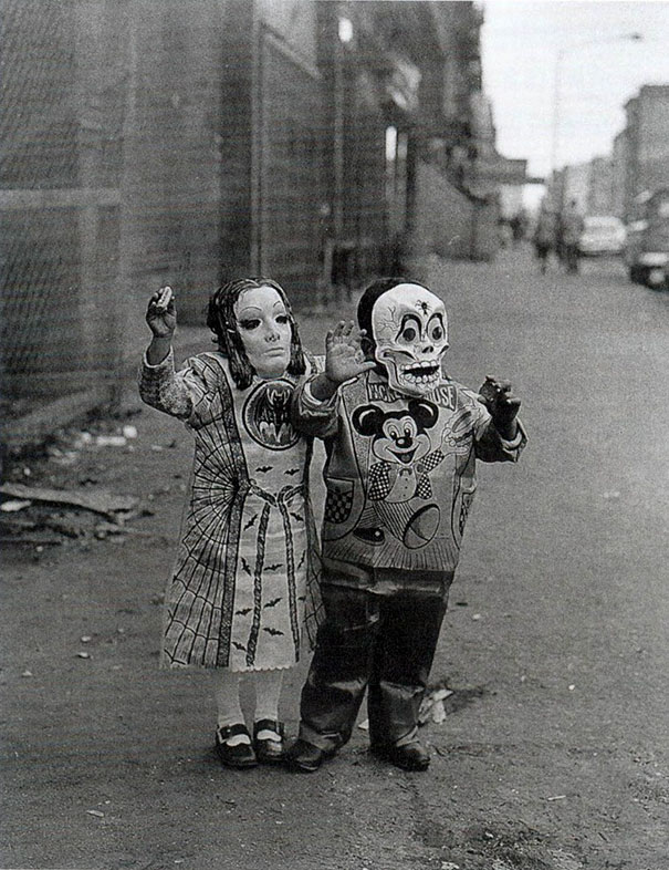 Two children wearing vintage Halloween costumes and masks, walking on a city street during Halloween festivities.