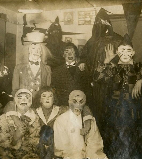 Group of people in vintage Halloween costumes with scary masks and outfits in a dimly lit room from early 20th century.