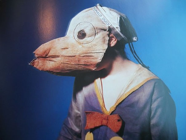 Person wearing a vintage Halloween costume with a large duck-like paper mask and a retro outfit against a blue background
