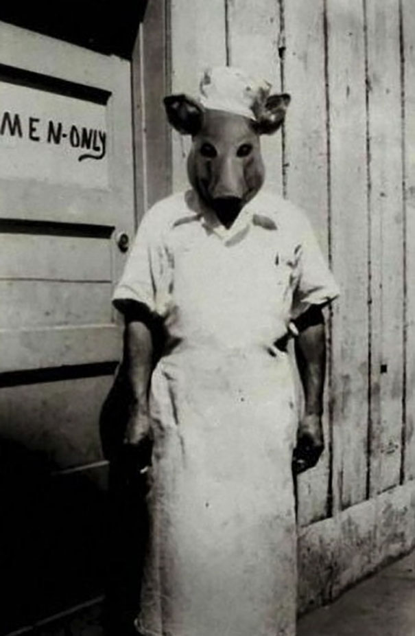 Person in vintage Halloween costume wearing a pig mask and chef outfit standing outside a men-only door in black and white photo
