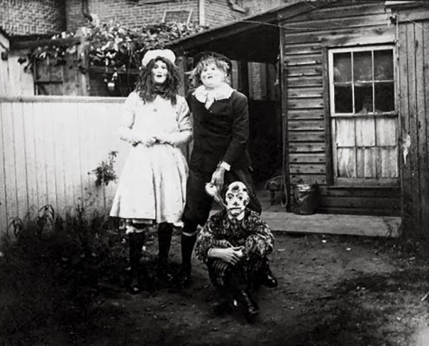 Three children in vintage Halloween costumes posing outside a rustic wooden house in a black and white photo.