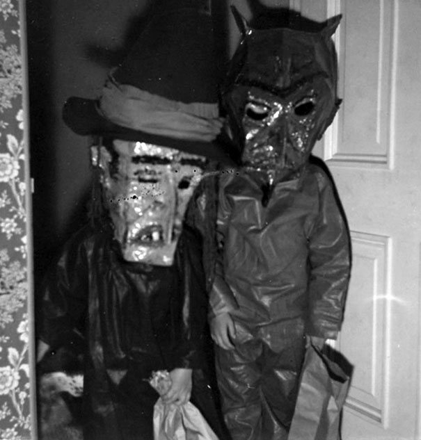 Two children in vintage Halloween costumes with homemade scary masks holding treat bags indoors.