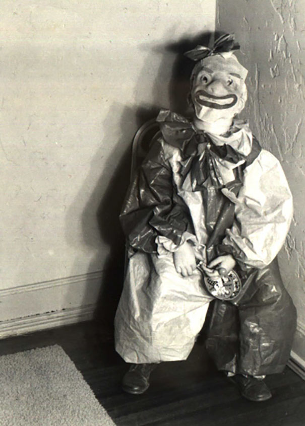 Vintage Halloween costume of a child dressed as a creepy clown sitting in a corner holding a small clock.