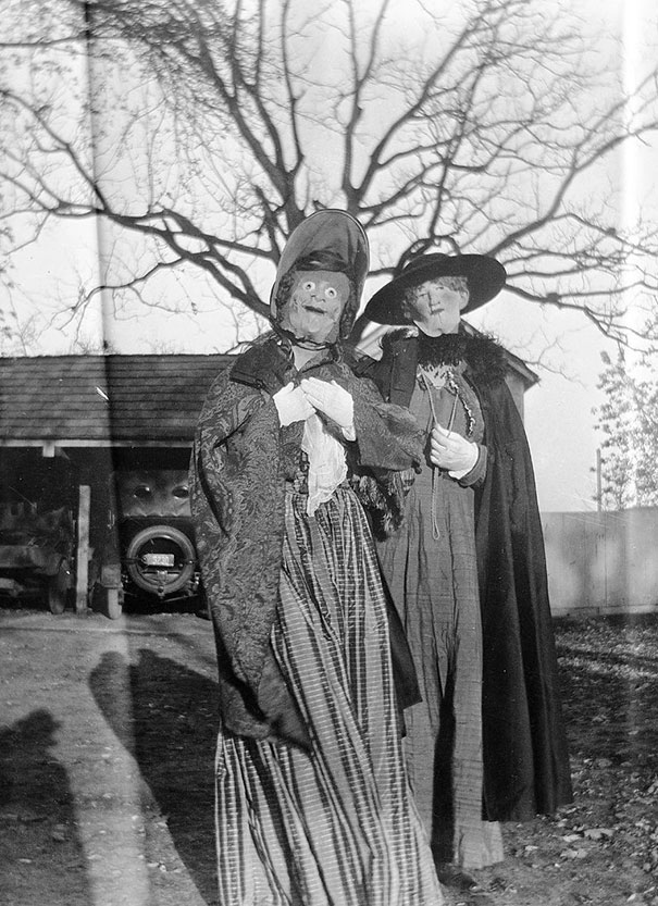 Two people wearing vintage Halloween costumes with eerie masks and old-fashioned dresses outdoors near a tree.