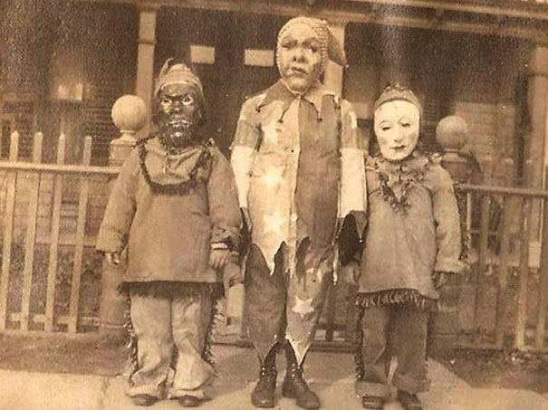 Three children in vintage Halloween costumes with eerie masks standing in front of a porch in an old photograph.