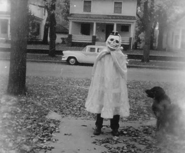 Child in vintage Halloween costume wearing skull mask and white dress standing on sidewalk with a dog nearby and a classic car in background.