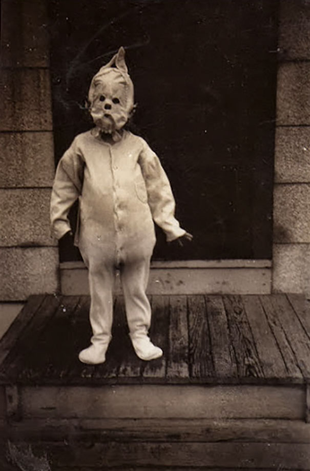 Child wearing a vintage Halloween costume with a spooky mask standing on a wooden porch in black and white.