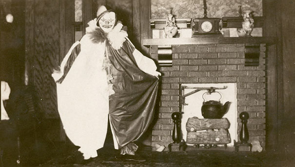 Vintage Halloween costume featuring a creepy clown with a large ruffled collar posing next to a brick fireplace indoors.