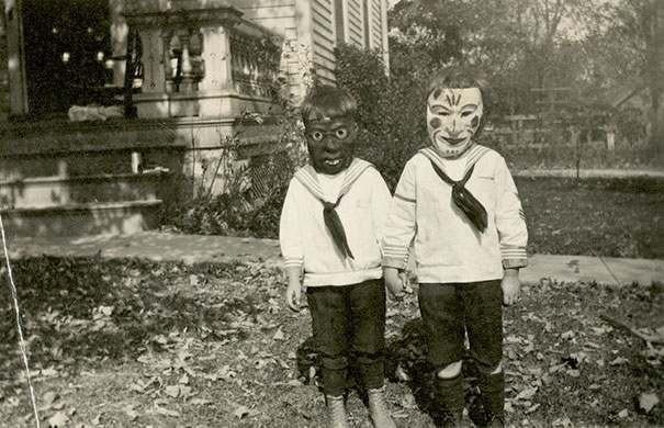 Two children wearing vintage Halloween costumes and masks, standing outside a house in an autumn setting.