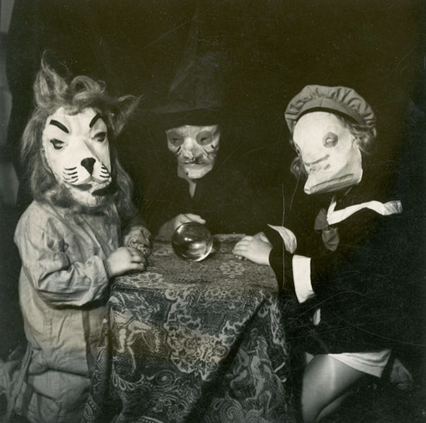 Three children in vintage Halloween costumes wearing homemade animal and ghost masks around a table with a crystal ball.