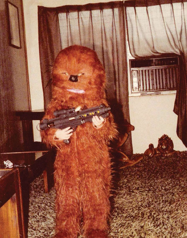Child in a vintage Halloween costume resembling a furry creature holding a toy gun indoors on a patterned carpet.