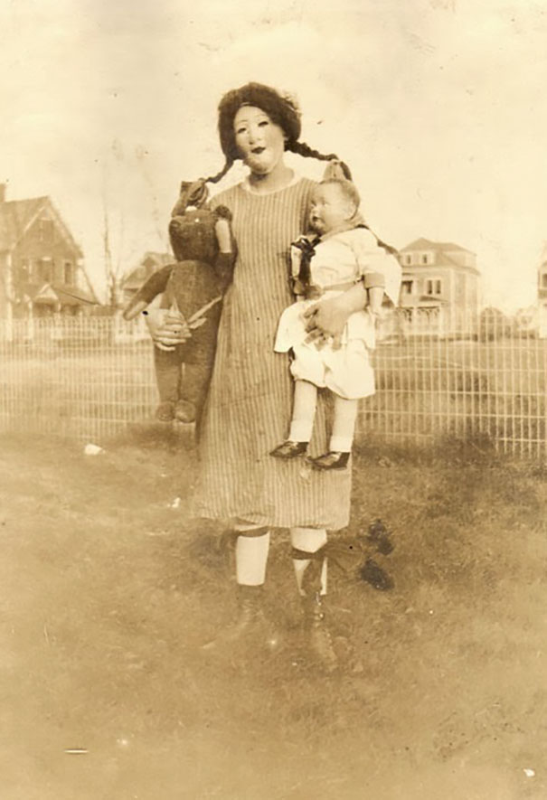 Woman in a vintage Halloween costume holding a baby and a large stuffed toy outside in a residential yard.