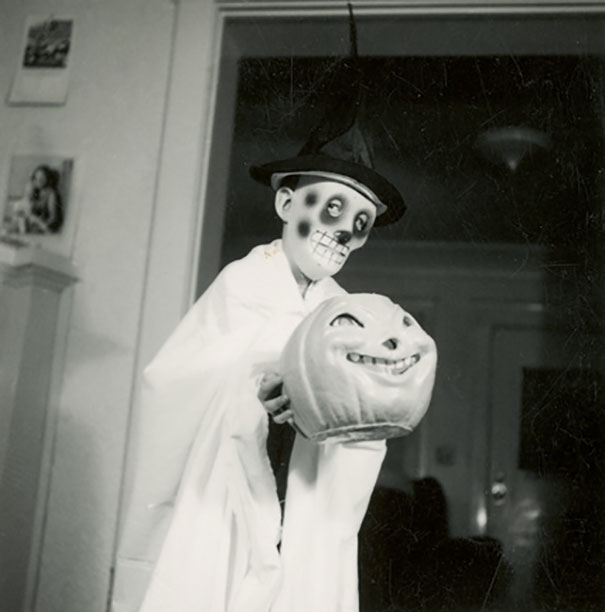 Child in vintage Halloween costume with skull makeup and witch hat holding a spooky pumpkin decoration indoors.