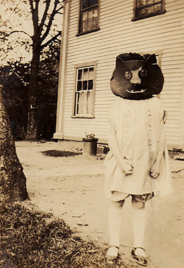 Child in a vintage Halloween costume with a large black cat mask standing outside an old house.