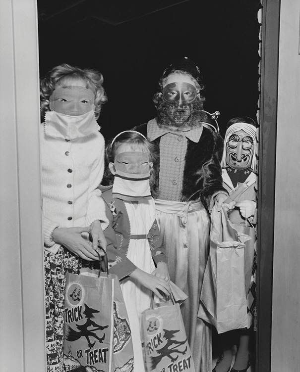 Four children wearing vintage Halloween costumes and masks, holding trick-or-treat bags at a doorway.