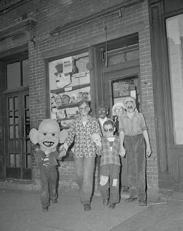 Group of children in vintage Halloween costumes wearing scary masks outside a brick building at night.